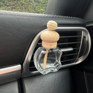 Puede incluir: Un ambientador de coche de vidrio transparente con una tapa de madera y un palo de madera en su interior. El ambientador está colgado de una ventilación de aire del coche.