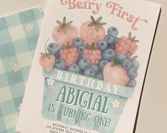 May include: A birthday invitation with a watercolor illustration of berries in a basket. The text "Berry First" and "Abigial is turning one!" are displayed in a whimsical font. The invitation is paired with a light blue gingham patterned envelope.
