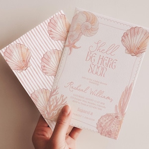 May include: Two baby shower invitations with a seashell theme. One invitation features a striped pattern with seashell illustrations. The other invitation has the text "Shell Be Here Soon" and additional details. The invitations are in shades of pink and peach.