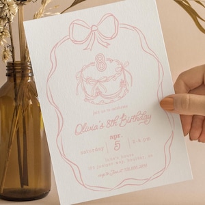 May include: A light pink birthday invitation with a bow and cake illustration. The invitation reads "Olivia's 8th Birthday" with event details. The invitation is held in front of a neutral background with a brown glass vase and dried plants.