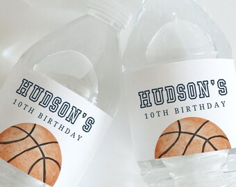May include: Two clear plastic water bottles with labels. The labels read "HUDSON'S 10TH BIRTHDAY" above a watercolour basketball design. The bottles are filled with water and have white caps.