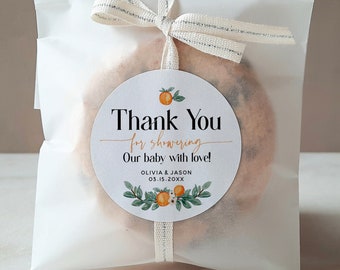 Op de afbeelding: Een witte cadeauzak met een wit lint eromheen gebonden. De zak bevat een ronde koek met een witte sticker waarop staat "Bedankt voor het douchen van onze baby met liefde! Olivia & Jason 03.15.202X". De sticker heeft een klein oranje bloemetjesontwerp.