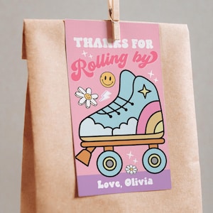 Może przedstawiać: Różowa etykieta prezentowa z ilustracją retro rolek i napisem "Thanks for Rolling by". Etykieta zawiera również emotikon, kwiat i imię "Olivia". Jest przymocowana do brązowej papierowej torby za pomocą drewnianego klipsa.