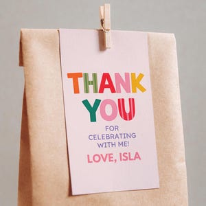 May include: A brown paper gift bag with a pink tag that says "THANK YOU" in colorful letters. The tag is clipped to the bag with a wooden clothespin. The text on the tag also reads "FOR CELEBRATING WITH ME! LOVE, ISLA."