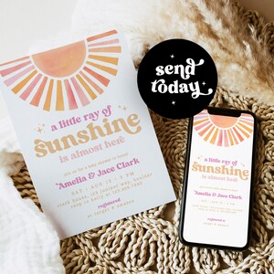 Peut inclure: Une invitation de f&ecirc;te pr&eacute;natale avec un design de coucher de soleil aquarelle. L'invitation indique "un petit rayon de soleil arrive bient&ocirc;t" et comprend les noms "Amelia & Jace Clark".
