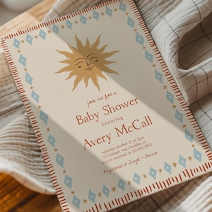 Op de afbeelding: Een rechthoekige uitnodigingskaart voor een babyshower met een crèmekleurige achtergrond, een gouden zonne-illustratie en een rand van blauwe ruiten en rode streepjes. De tekst luidt: join us for a Baby Shower honoring Avery McCall, saturday, october 10 at 2pm, 123 juniper way, boulder, co, 123.456.7890, rsvp to courtney at 123.456.7890, Registered at Target + Amazon.