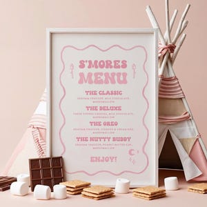 以下が含まれることがあります： 背景にティピーがある、ピンクと白の額入りの「S'Mores Menu」サイン。メニューには、スモアのバリエーションが記載されています。マシュマロ、グラハムクラッカー、チョコレートバーが看板の前に配置されています。