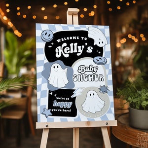 May include: A blue and white checkered sign with a black circle in the center. The sign says "Welcome to Kelly's Baby Shower" with a ghost and smiley face graphic. The sign is on a wooden easel.