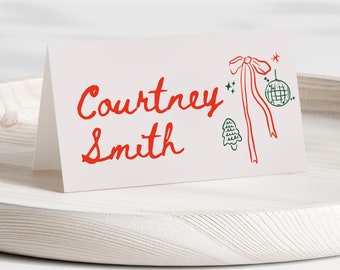 Puede incluir: Una tarjeta de lugar blanca con texto rojo en cursiva que dice "Courtney Smith". La tarjeta está decorada con un lazo rojo, un árbol de Navidad verde y una bola de discoteca verde y blanca.