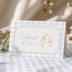 May include: A folded card with a blue gingham pattern and a white central panel. The panel features the words "Thank You" in script, along with illustrations of two geese with blue bows. Daisies and other small white flowers are in the background.
