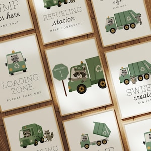 May include: A collection of framed prints with a green garbage truck and raccoon illustrations. The prints feature phrases like "Dump Gifts Here," "Refueling Station," and "Loading Zone." The signs are on a wooden surface.