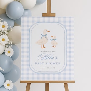 May include: A baby shower sign on a wooden easel. The sign has a blue and white gingham background with a framed illustration of two ducks. The text reads "-welcome to- Nila's BABY SHOWER August 13, 20XX". Balloons and flowers are in the background.