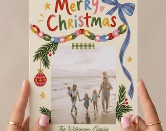 Peut inclure: Une carte de Noël rectangulaire avec le texte Merry Christmas en lettres colorées, un ruban bleu, une guirlande de chaînes en papier et des branches de pin. Une photo centrale montre une famille sur une plage, avec le texte The Wilkinson Family 2026.