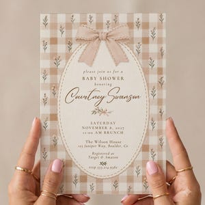May include: A rectangular baby shower invitation card featuring a beige and white gingham pattern with small floral sprigs. A central oval frame contains a beige bow and text: please join us for a BABY SHOWER honoring Courtney Swanson.