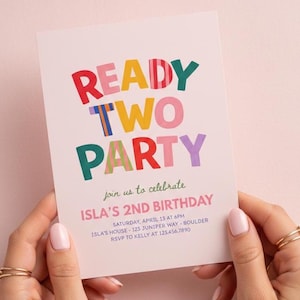 以下が含まれることがあります： カラフルなブロック文字で「READY TWO PARTY」と書かれた白い招待状。 イスラの2歳の誕生日のお祝いを、日付、時間、場所を含めて発表しています。 金色の指輪をした手でカードが持たれています。
