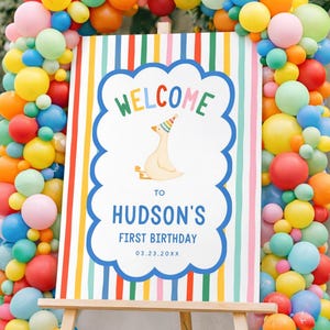 May include: A colorful birthday party sign on an easel, framed by a vibrant balloon arch. The sign reads "WELCOME to HUDSON'S FIRST BIRTHDAY 03.23.20XX" with a cartoon goose wearing a party hat. The balloons are in shades of red, yellow, blue, orange, and pink.