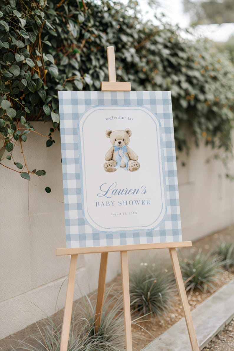 May include: A baby shower sign on a wooden easel. The sign has a light blue gingham pattern border and features a teddy bear illustration with a blue ribbon. Text reads "Welcome to Lauren's Baby Shower" and the date.