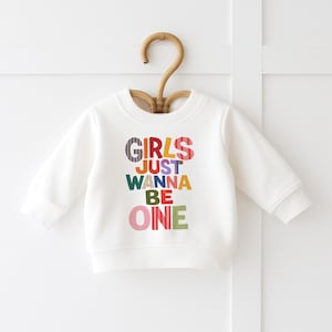 May include: A white long-sleeve crewneck sweatshirt featuring the colorful text "GIRLS JUST WANNA BE ONE" printed on the front. The letters are designed in various patterns and solid colors, including stripes, pink, yellow, green, and blue.