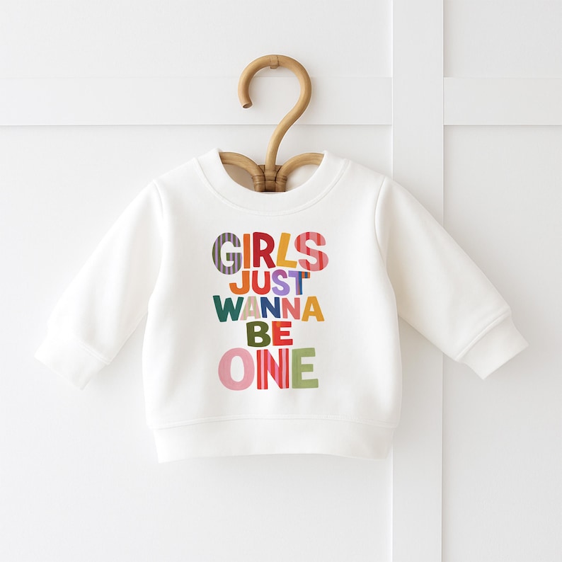 May include: A white long-sleeve crewneck sweatshirt featuring the colorful text "GIRLS JUST WANNA BE ONE" printed on the front. The letters are designed in various patterns and solid colors, including stripes, pink, yellow, green, and blue.