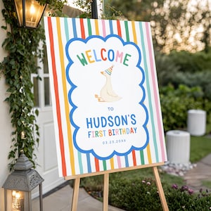 Puede incluir: Un letrero de bienvenida rectangular con un centro blanco y borde festoneado azul, una oca de dibujos animados con un gorro de fiesta y el texto "WELCOME TO HUDSON'S FIRST BIRTHDAY 03.23.20XX". El fondo tiene rayas verticales de colores arco&iacute;ris.