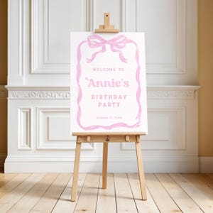 May include: A pink and white birthday party welcome sign on a wooden easel. The sign reads "Welcome to Annie's Birthday Party" with a pink bow and ribbon design. The date "October 13, XXXX" is at the bottom.