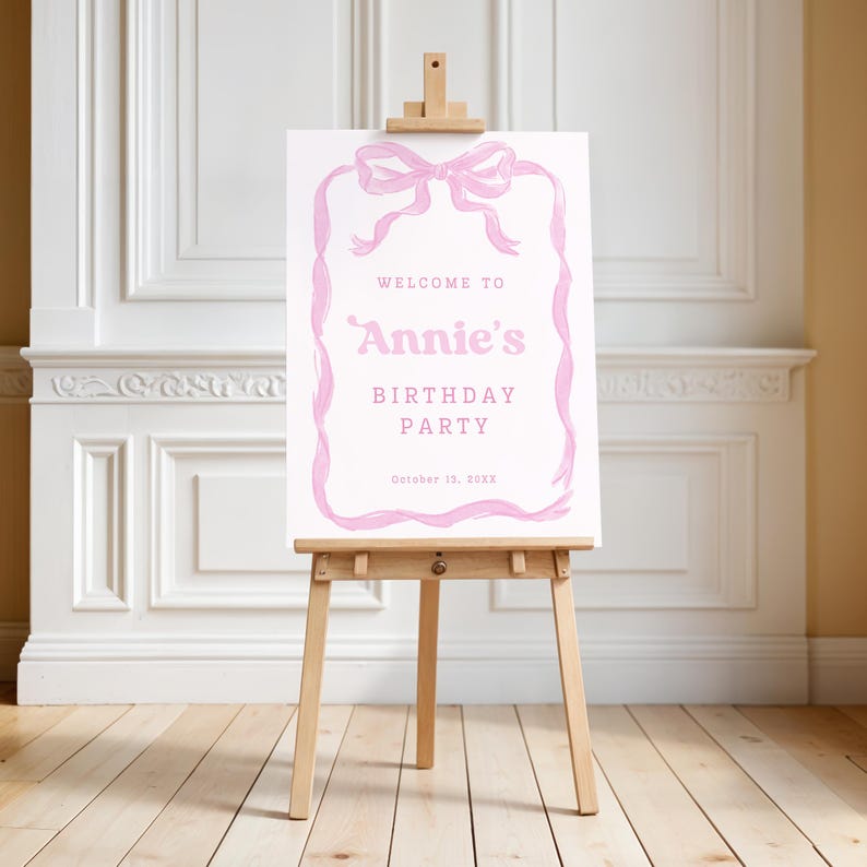 May include: A pink and white birthday party welcome sign on a wooden easel. The sign reads "Welcome to Annie's Birthday Party" with a pink bow and ribbon design. The date "October 13, XXXX" is at the bottom.