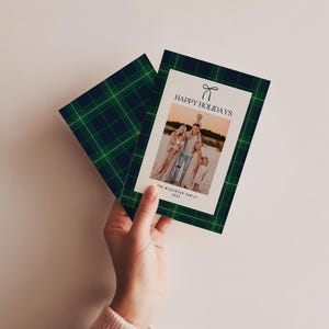 May include: A holiday card with a family photo, featuring a green and navy plaid pattern border. The card reads "HAPPY HOLIDAYS" with a bow graphic. The family photo is on a white background.