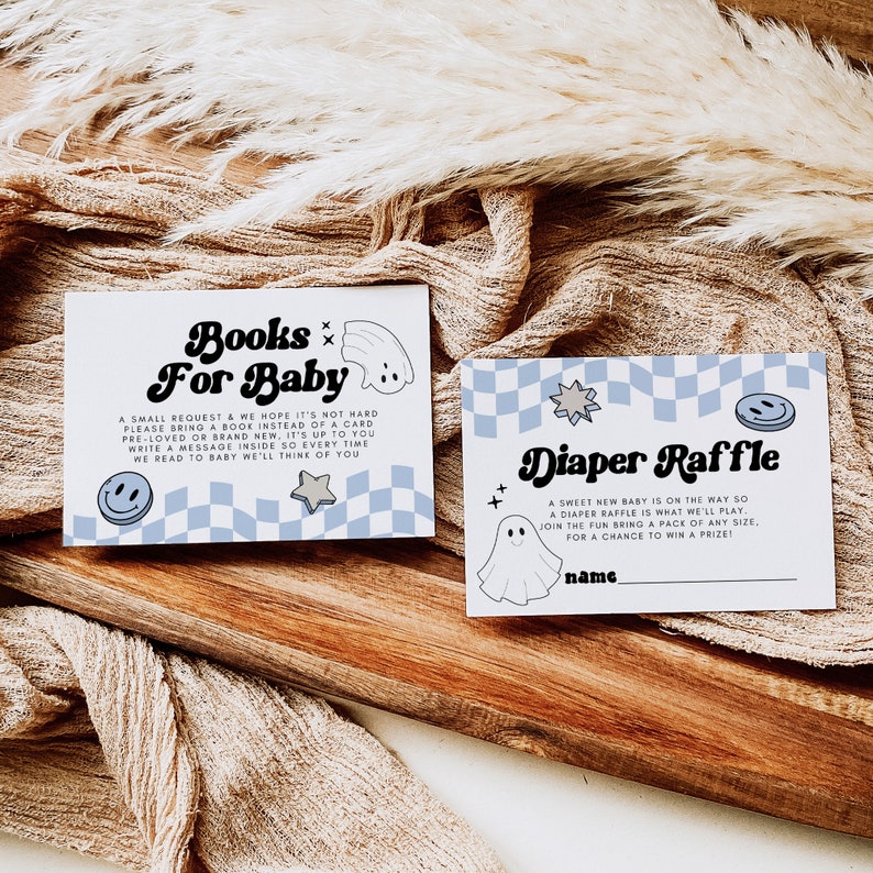 May include: Two baby shower cards on a wooden surface. One card reads "Books For Baby" with a ghost and smiley face graphic. The other card reads "Diaper Raffle" with a ghost and star graphic. Both cards have a blue and white checkered pattern.