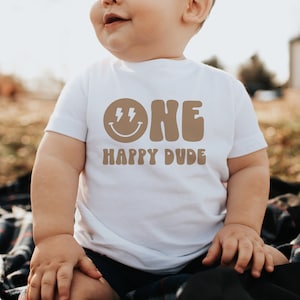 May include: A white t-shirt with a brown graphic that says "ONE" in a retro font with a smiley face and lightning bolts. Below the text is the phrase "HAPPY DUDE".