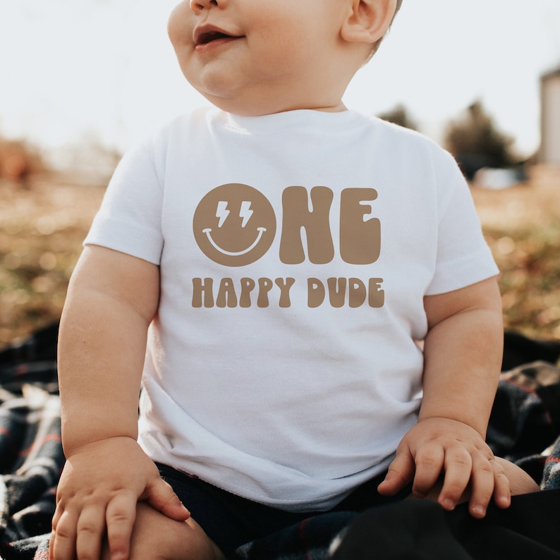 May include: A white t-shirt with a brown graphic that says "ONE" in a retro font with a smiley face and lightning bolts. Below the text is the phrase "HAPPY DUDE".