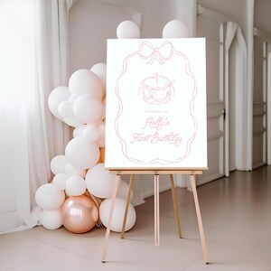 May include: A white sign on a wooden easel with a pink outline of a cake and bow. The sign reads "welcome to Holly's First Birthday" in pink script. White and rose gold balloons are arranged to the left of the sign.