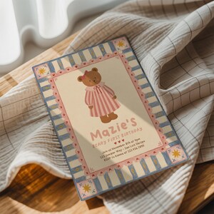 May include: A rectangular birthday invitation card featuring a brown teddy bear in a pink striped dress. The border has blue and white stripes with pink scalloped edges and star accents. Text reads: Mazie's BEARY FIRST BIRTHDAY, November 8th at 4pm, 123 Juniper Way, San Luis Obispo, RSVP to Kenna at 123 456 7890.