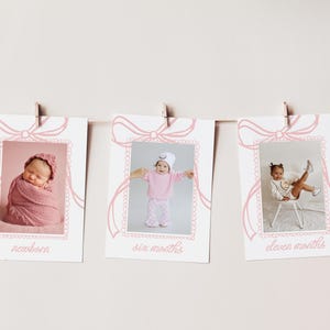 May include: A set of three baby milestone photo cards hanging on a string. Each card features a pink bow design and a photo of a baby at different ages: newborn, six months, and eleven months. The cards are printed with the age of the baby.