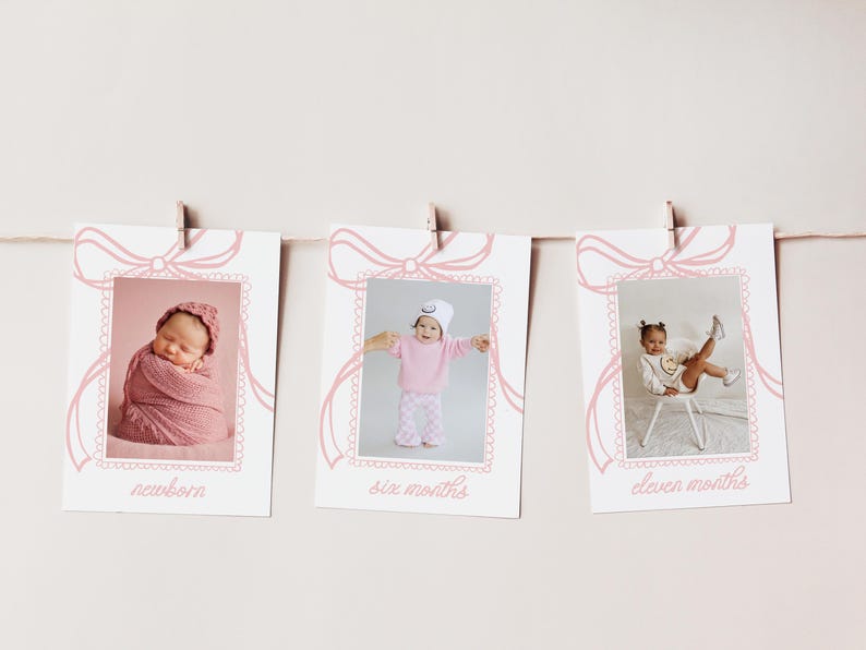 May include: A set of three baby milestone photo cards hanging on a string. Each card features a pink bow design and a photo of a baby at different ages: newborn, six months, and eleven months. The cards are printed with the age of the baby.