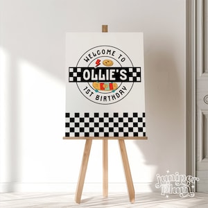 May include: A white sign with a black and white checkered border. The sign has a black circle with the text "Welcome to Ollie's 1st Birthday" and a skateboard graphic inside the circle.
