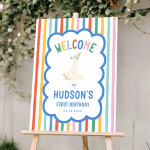 May include: A colorful welcome sign for Hudson's first birthday, displayed on a wooden easel. The sign features a cartoon goose wearing a party hat, the word "WELCOME" in rainbow colors, and the date 03.23.20XX.
