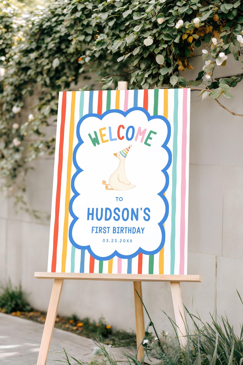 May include: A colorful welcome sign for Hudson's first birthday, displayed on a wooden easel. The sign features a cartoon goose wearing a party hat, the word "WELCOME" in rainbow colors, and the date 03.23.20XX.