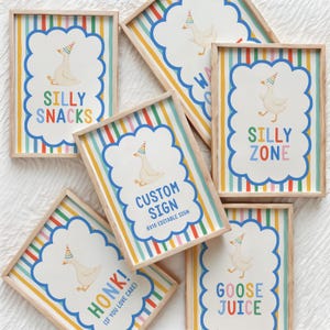 Puede incluir: Cinco marcos rectangulares de madera con impresiones sobre un fondo de rayas verticales de arco&iacute;ris. Cada impresi&oacute;n presenta un borde festoneado blanco, un ganso de dibujos animados con un gorro de fiesta y texto que incluye SILLY SNACKS, SILLY ZONE, CUSTOM SIGN 8X10, HONK! (IF YOU LOVE CAKE) y GOOSE JUICE.