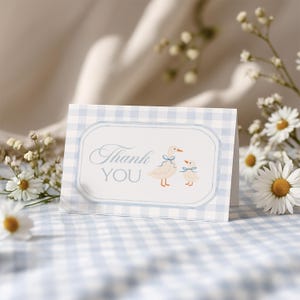 May include: A folded card with a blue gingham pattern and a white central panel. The panel features the words "Thank You" and illustrations of two geese with blue bows. Daisies and other small white flowers are in the background.