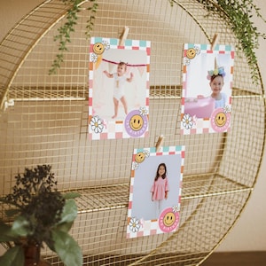 May include: Three pink and white photo cards with a checkered pattern and a smiley face design. Each card has a photo of a child. The cards are hanging on a gold wire rack.
