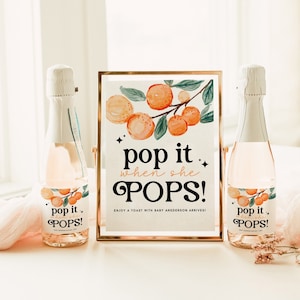 May include: A gold framed sign with a watercolor illustration of orange blossoms and the text "pop it when she POPS! Enjoy a toast with baby Anderson arrives!" Two champagne bottles with labels that say "pop it when she POPS!"