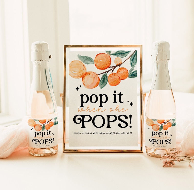 May include: A gold framed sign with a watercolor illustration of orange blossoms and the text "pop it when she POPS! Enjoy a toast with baby Anderson arrives!" Two champagne bottles with labels that say "pop it when she POPS!"