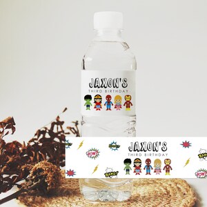 May include: Water bottle label with a superhero theme. The label features a white background with colorful cartoon superheroes, the text "Jaxon's Third Birthday", and comic book sound effects like "Pow!" and "Bang!"