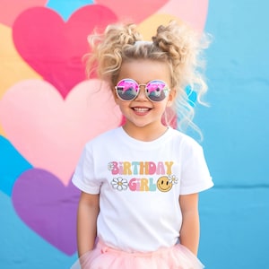 May include: A white t-shirt with a colorful "Birthday Girl" graphic featuring daisies and a smiley face. The shirt is worn by a young girl with blonde hair and pink sunglasses.