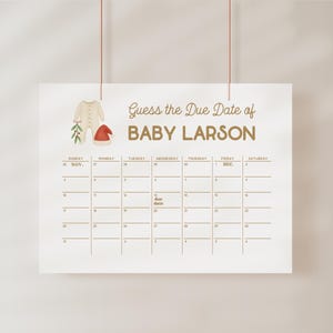 May include: A white paper calendar with the text "Guess the Due Date of BABY LARSON" in gold lettering. The calendar has a grid of dates and illustrations of baby clothes. The calendar is hanging from two red strings.