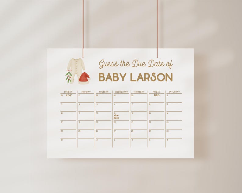 May include: A white paper calendar with the text "Guess the Due Date of BABY LARSON" in gold lettering. The calendar has a grid of dates and illustrations of baby clothes. The calendar is hanging from two red strings.