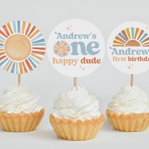 May include: Three cupcakes with white frosting, each topped with a decorative circle. The circles feature a sun design and text that reads "Andrew's one happy dude" and "Andrew's first birthday". The color scheme includes orange, blue, and beige.
