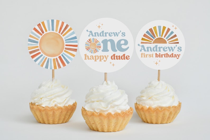 May include: Three cupcakes with white frosting, each topped with a decorative circle. The circles feature a sun design and text that reads "Andrew's one happy dude" and "Andrew's first birthday". The color scheme includes orange, blue, and beige.