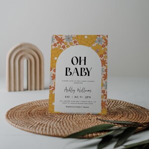 May include: A baby shower invitation with a floral pattern in shades of yellow, orange, and blue. The text reads "Oh Baby" and includes the name "Ashley Williams", the date "Sat Jul 9 1 PM", and the address "125 Juniper Plum Way, Santa Cruz, CA". The invitation also includes the RSVP information and the registry information.
