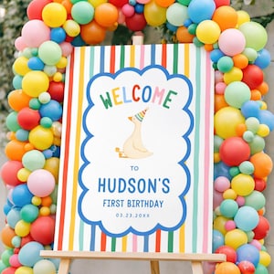 Puede incluir: Un letrero de cumplea&ntilde;os rectangular sobre un caballete de madera rodeado por un arco de globos de colores. El letrero presenta rayas verticales, un borde azul festoneado, un pato de dibujos animados con gorro de fiesta y el texto WELCOME TO HUDSON'S FIRST BIRTHDAY 03.23.20XX.